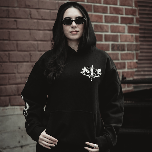 A woman in the Kittie Hecate - Black Pullover, a heavyweight fleece with an oversized fit, stands confidently with long dark hair and black sunglasses against a red brick wall.
