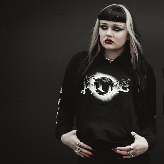 A woman with black and white hair, heavy eyeliner, and dark lipstick stands against a dark background with a serious expression, wearing the Kittie Nox - Black Cropped Women's Pullover with her hands in her pockets.