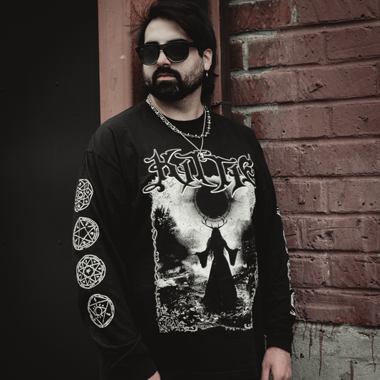 A man with dark hair, a beard, and sunglasses stands by a brick wall wearing the Kittie Eclipse - Black Long Sleeve, garment-dyed and made from ring spun cotton, featuring occult sleeve symbols and an artistic horned figure on the front.