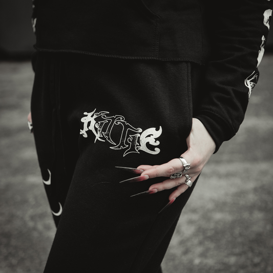 A person with long silver nails and rings rests their hand on relaxed fit black Hecate Sweatpants by Kittie, featuring ornate white gothic lettering—an ideal bold statement for a unique streetwear look.