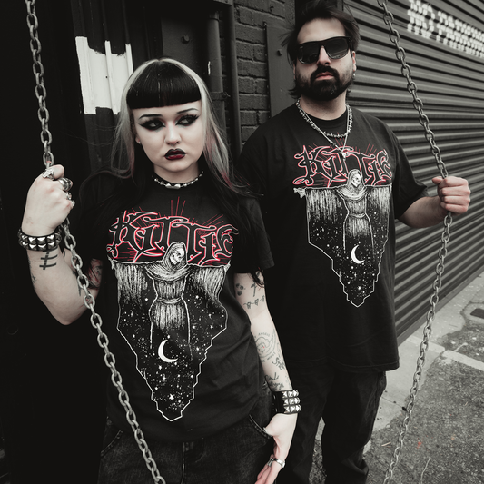Two people dressed in black gothic style stand by a metal gate, both wearing matching Kittie Reaper Black T-Shirts made of soft ring-spun cotton. They have dark makeup, piercings, tattoos, and chains draped around them.